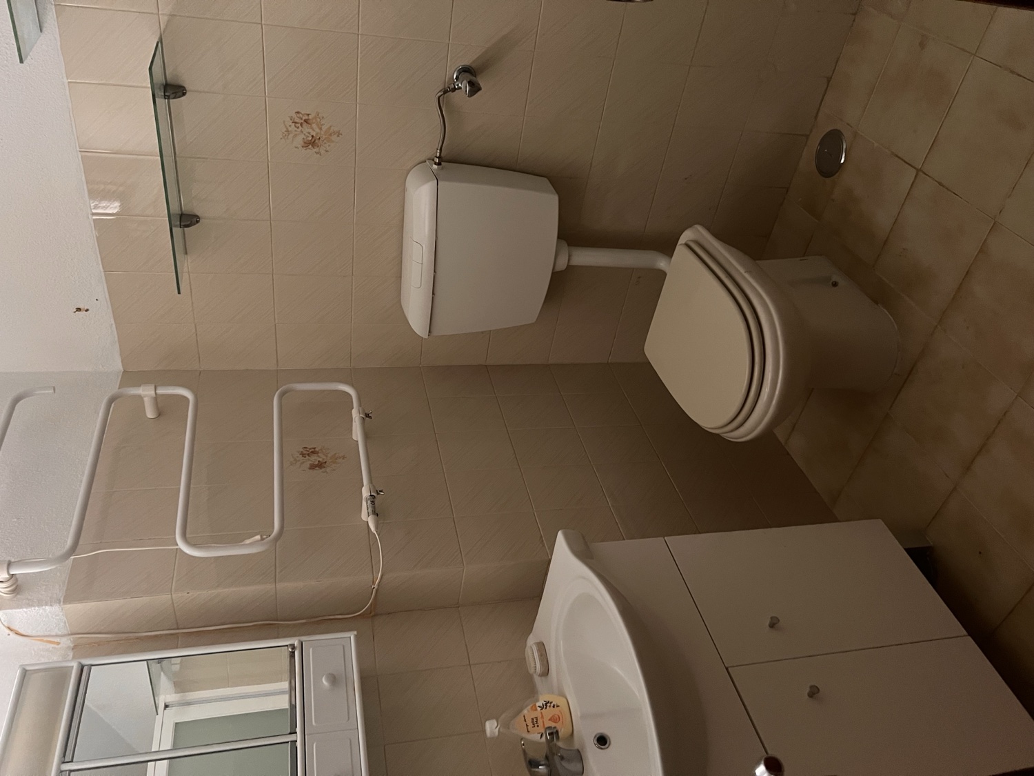 Vista before &mdash; cream-tiled bathroom with white toilet, wall-mounted sink, glass shower screen, and a towel-rail radiator